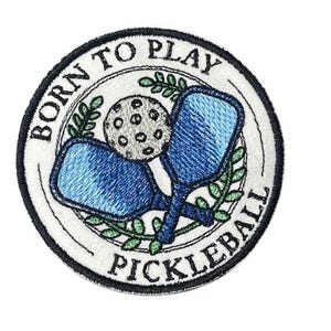 May include: Embroidered patch with the text "BORN TO PLAY PICKLEBALL." The patch features two blue pickleball paddles crossed over a pickleball ball. Green leaves and a white background complete the design.