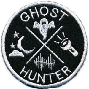 May include: A black and white embroidered patch with the words "Ghost Hunter" in a circle. The patch features a ghost, a crescent moon, a flashlight, and a series of lines that resemble sound waves.