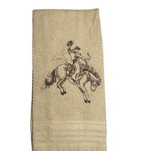 Op de afbeelding: Een beige keukendoek met een bruin geborduurd ontwerp van een cowboy die een bokkend paard berijdt.