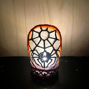 May include: A Halloween candle holder featuring a spider web and spider design. The candle holder is orange and white with a black spider web and spider design. The candle holder is lit from within, creating a spooky glow.