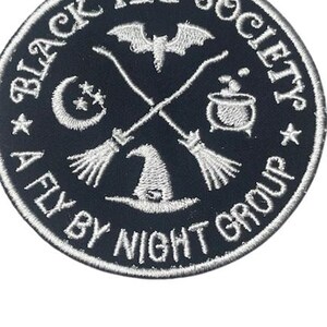 Op de afbeelding: Ronde zwarte patch met wit geborduurde tekst en afbeeldingen. De tekst luidt "BLACK HAT SOCIETY" en "A FLY BY NIGHT GROUP". De afbeeldingen omvatten een vleermuis, gekruiste bezems, een heksenhoed, een maan, sterren en een borrelende ketel.