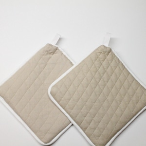 Puede incluir: Dos agarradores de cocina cuadrados de color beige con un patrón de diamantes acolchado y ribete de tela blanca en los bordes. Cada agarrador incluye una pequeña trabilla de tela blanca en la esquina superior para colgarlo, un accesorio práctico.