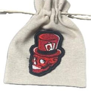 Könnte beinhalten: Ein kleiner, beigefarbener Kordelzugbeutel mit einem roten Totenkopf-Aufnäher. Der Aufnäher zeigt einen Totenkopf mit Zylinder und Spielkarten. Der Beutel ist aus einem weichen, strukturierten Stoff gefertigt und hat einen weißen Kordelzugverschluss.