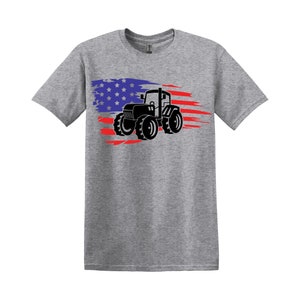 Può includere: Una maglietta grigia con una silhouette nera di un trattore che guida attraverso un design di bandiera americana.