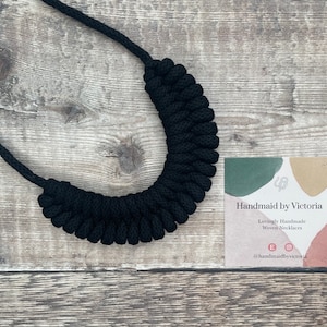 May include: Black woven rope necklace on a wooden surface. The necklace is handmade and is a statement piece. The tag reads "Handmade by Victoria" and "Lovingly Handmade Woven Necklaces".