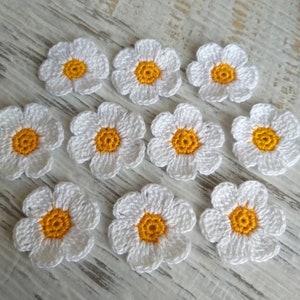 May include: Ten white crocheted daisy flowers with yellow centers. The flowers are arranged in a semi-circle on a light wood surface.