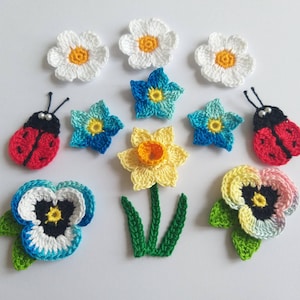 May include: A collection of crochet appliques featuring three white flowers with yellow centres, two red ladybirds with black heads and white bead eyes, four blue flowers, one yellow daffodil with a stem and leaves, and two pansies.
