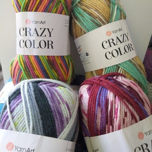 May include: Four skeins of YarnArt Crazy Color yarn in various color combinations. The skeins are labeled with the brand name and product name. The yarn colors include green, yellow, pink, purple, and blue. The yarn is tightly wound.