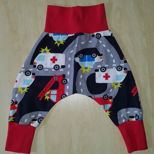 Könnte beinhalten: Rote und schwarze Babyhose mit einem Aufdruck von Einsatzfahrzeugen, darunter Krankenwagen, Polizeiautos und Feuerwehrautos. Die Hose hat einen roten elastischen Bund und rote Bündchen.
