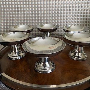 Op de afbeelding: Zes zilverkleurige serveerschalen met voetstukken, geplaatst op een donkerhouten dienblad met een zilveren rand. Elke schaal heeft een ondiepe, ronde vorm en een korte, decoratieve voet. De achtergrond is een patroonstof.