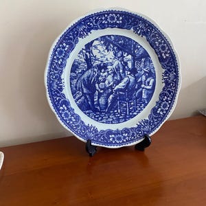 Könnte beinhalten: Blau-weiße dekorative Platte mit einer detaillierten Szene und floralem Rand. Die Platte wird auf einem schwarzen Ständer präsentiert, der ihr aufwendiges Design und ihre Vintage-Ästhetik hervorhebt. Der Rand der Platte hat einen gewellten Rand.