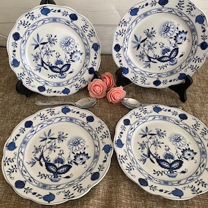 Peut inclure: Quatre assiettes en céramique blanche avec un motif floral bleu et des bords festonnés. Chaque assiette présente un motif floral central et une bordure avec des feuilles et des baies stylisées. Les assiettes sont présentées sur des supports noirs.