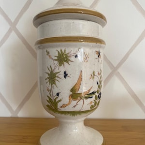 May include: A cream ceramic jar with a lid, decorated with a painted bird, foliage, and butterflies. The jar has a pedestal base and brown trim. The jar is approximately 25 cm tall.