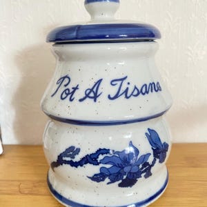 Può includere: Vaso in ceramica bianca con coperchio e base blu, con la scritta "Pot A Tisane" in blu. Il vaso è decorato con disegni floreali blu e macchie.