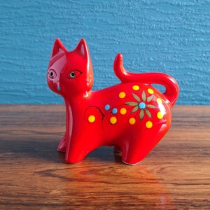 May include: A bright red ceramic cat ornament with a glossy finish. The cat has a whimsical design with yellow and blue polka dots, a floral motif, and a curled tail. The figurine is standing on four legs and has a detailed face with green eyes.