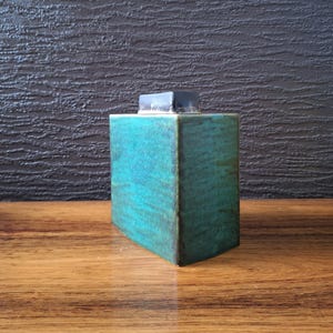 May include: A square, teal ceramic container with a dark grey lid. The container has a textured surface and sits on a wooden surface. The teal colour has darker green and brown accents.
