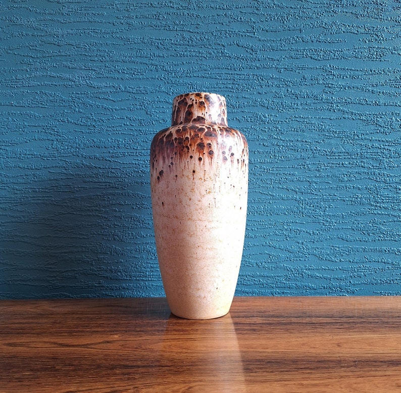 May include: A white ceramic vase with brown speckles and drips. The vase has a rounded bottom and a narrow opening.