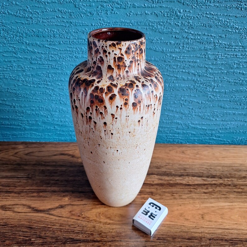 May include: A white ceramic vase with brown speckles and a drip glaze effect. The vase is tall and narrow with a rounded bottom and a slightly flared opening.