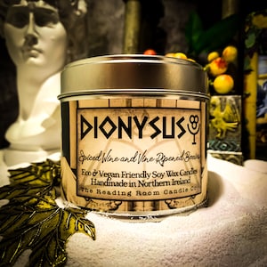 May include: A silver-lidded candle tin with a label that reads "DIONYSUS" in black lettering. The label also states "Spiced Wine and Vine Ripened Berries" and "Eco & Vegan Friendly Soy Wax Candles". The Reading Room Candle Co.