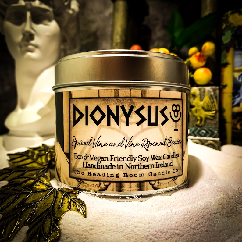 May include: A silver-lidded candle tin with a label that reads "DIONYSUS" in black lettering. The label also states "Spiced Wine and Vine Ripened Berries" and "Eco & Vegan Friendly Soy Wax Candles". The Reading Room Candle Co.