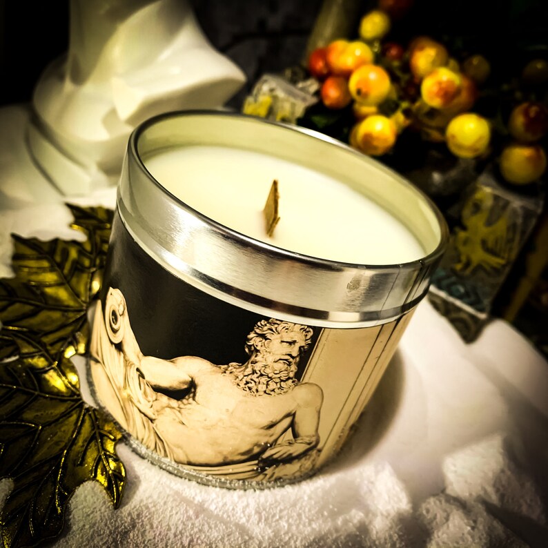 May include: A scented candle in a black and silver container. The candle has a white wax and a wooden wick. The container features a classical illustration of a reclining figure. The candle sits on a gold leaf.