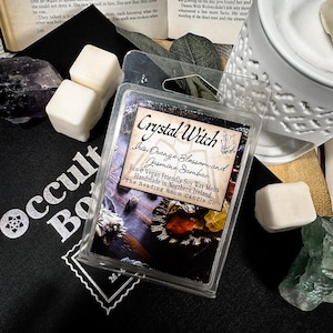 Puede incluir: Un paquete de plástico transparente de ceras de soja Crystal Witch. La etiqueta dice "Iris, Flor de Naranja y Jazmín Sambac". Dos cubos de cera blancos están junto al paquete, junto con una tela negra con "Occult Books" impreso.