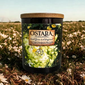 May include: A dark candle with a wooden lid, labelled "OSTARA" with "Sweet Grass and Bergamot" text. The label also states "Eco & Vegan Friendly Soy Wax Candles Handmade in Northern Ireland". The candle is set against a field of flowers.