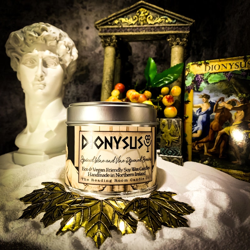 May include: A round, silver-lidded candle tin with the word "DIONYSUS" in bold letters. The label reads "Spiced Wine and Vine Ripened Berries" and "Eco & Vegan Friendly Soy Wax Candles." The candle sits on a bed of white material with gold leaf accents.