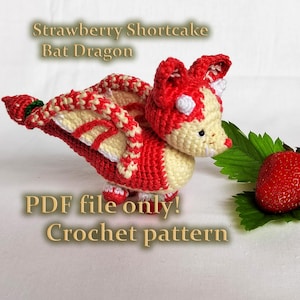 May include: Crochet pattern for a red and cream coloured bat dragon amigurumi. The dragon is sitting on a white surface with a strawberry and green leaves in the background. The text "Strawberry Shortcake Bat Dragon" and "PDF file only! Crochet pattern" is visible in the image.