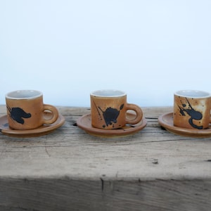 May include: Three brown ceramic coffee cups with black splatter paint designs, each sitting on a matching saucer.