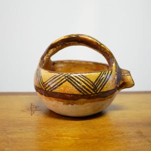 May include: A small, hand-crafted ceramic bowl with a handle. The bowl is decorated with a geometric pattern in brown and yellow paint.