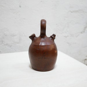 May include: A brown ceramic pitcher with two spouts and a handle. The pitcher has a handwritten letter "L" on the side.
