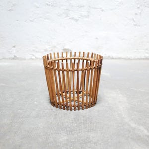 May include: A round, brown, woven bamboo planter with a natural finish. The planter is made of thin, vertical strips of bamboo that are woven together to create a cylindrical shape.