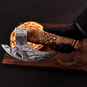 Op de afbeelding: Een Viking-stijl bijl met een houten handvat en een decoratief blad. Het blad heeft ge&euml;tste ontwerpen en het handvat is omwikkeld met touw. De bijl wordt boven een houten snijplank met een gebakken item gehouden.