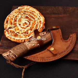 Op de afbeelding: Een kleine, decoratieve bijl met een houten handvat en een leren schede. De handgreep is omwikkeld met touw. De bijl staat naast een pizza met een witte saus swirl design. Het woord "ProAction" is zichtbaar.