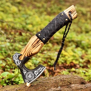 May include: A handcrafted axe with a wooden handle and a black, patterned leather grip. The axe head is black with intricate silver designs and a circular emblem. A leather strap is attached to the handle. The axe is resting on a log.
