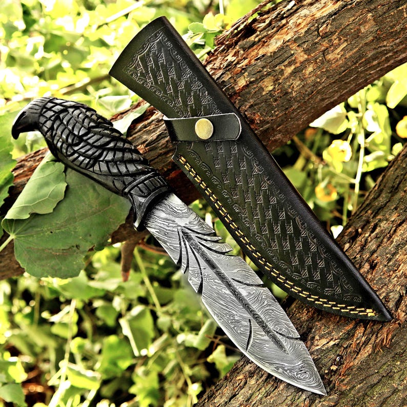 Puede incluir: Cuchillo de acero de Damasco con mango tallado con cabeza de &aacute;guila y funda de cuero negro. La hoja tiene un dise&ntilde;o de pluma. La funda est&aacute; grabada con un patr&oacute;n geom&eacute;trico y tiene costuras doradas.