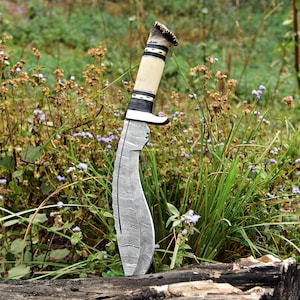 Puede incluir: Un cuchillo grande y curvo con una hoja estampada est&aacute; clavado en un trozo de madera. El mango es de color claro con detalles en negro y dorado y un extremo texturizado. El cuchillo est&aacute; rodeado de hierba verde y peque&ntilde;as flores moradas.