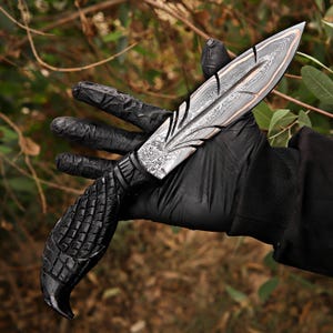 Puede incluir: Un cuchillo artesanal con una hoja estampada y un mango oscuro y texturizado. La hoja tiene una apariencia en capas con un borde color cobre. El mango est&aacute; intrincadamente tallado. El cuchillo se sostiene en una mano enguantada sobre un fondo natural borroso.