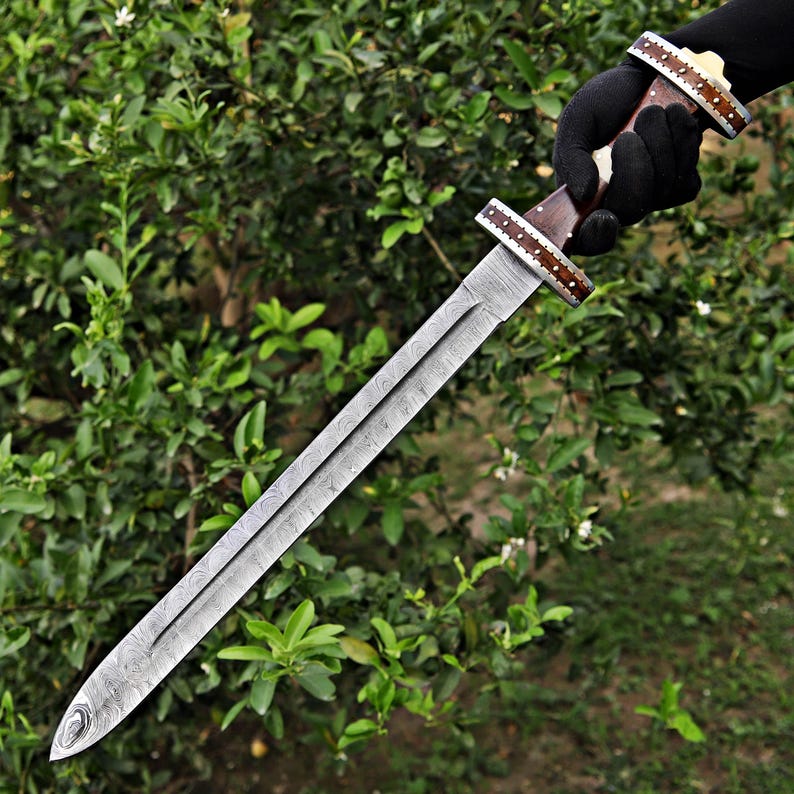 May include: A long, silver sword with a patterned blade and a wooden handle. The handle is adorned with decorative accents. The sword is held against a backdrop of green foliage.