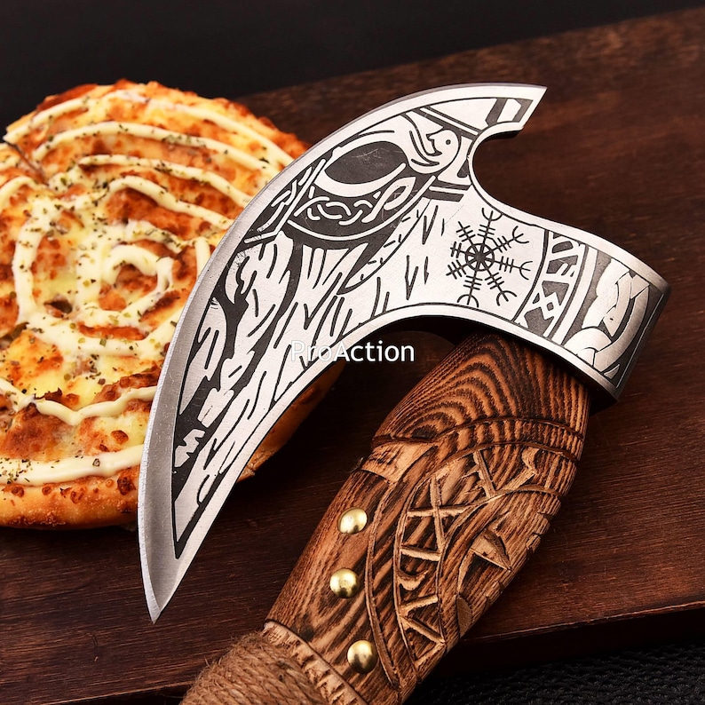 Op de afbeelding: Een Viking-stijl bijl met een gebogen blad en een houten handvat. Het blad heeft ge&euml;tste ontwerpen, waaronder een vogel en een kompassymbool. De handgreep is gesneden met ingewikkelde patronen en messing studs. De bijl wordt getoond op een houten oppervlak.