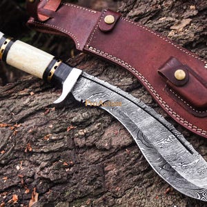 Puede incluir: Un cuchillo kukri artesanal con una hoja curva y un mango de hueso, acentuado con detalles negros y dorados. La hoja presenta un dise&ntilde;o &uacute;nico. Se incluye una funda de cuero marr&oacute;n con cierre de bot&oacute;n a presi&oacute;n.