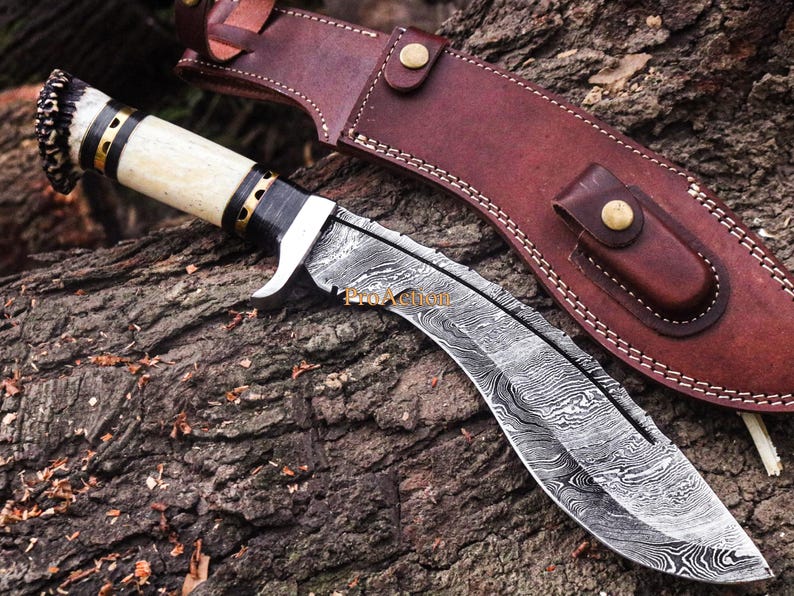 Puede incluir: Un cuchillo kukri artesanal con una hoja curva y un mango de hueso, acentuado con detalles negros y dorados. La hoja presenta un dise&ntilde;o &uacute;nico. Se incluye una funda de cuero marr&oacute;n con cierre de bot&oacute;n a presi&oacute;n.