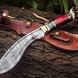 May include: A Damascus steel knife with a curved blade, red wooden handle, and a leather sheath. The handle has brass accents and a decorative end. The knife and sheath are resting on a textured wooden surface.