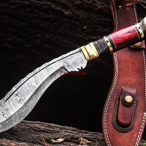 May include: A Damascus steel kukri knife with a curved blade and a handle featuring red wood, brass accents, and a textured antler end. The knife rests on a wooden surface next to a brown leather sheath.