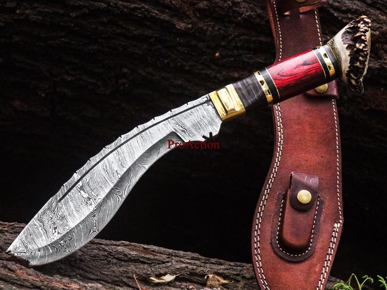 May include: A Damascus steel kukri knife with a curved blade and a handle featuring red wood, brass accents, and a textured antler end. The knife rests on a wooden surface next to a brown leather sheath.