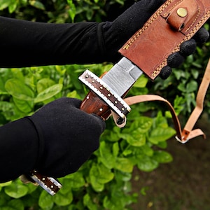 May include: A handcrafted knife with a patterned steel blade and a wooden handle. The handle is adorned with decorative metal accents. The knife is partially sheathed in a brown leather case with a strap. The background is a blurred view of green foliage.