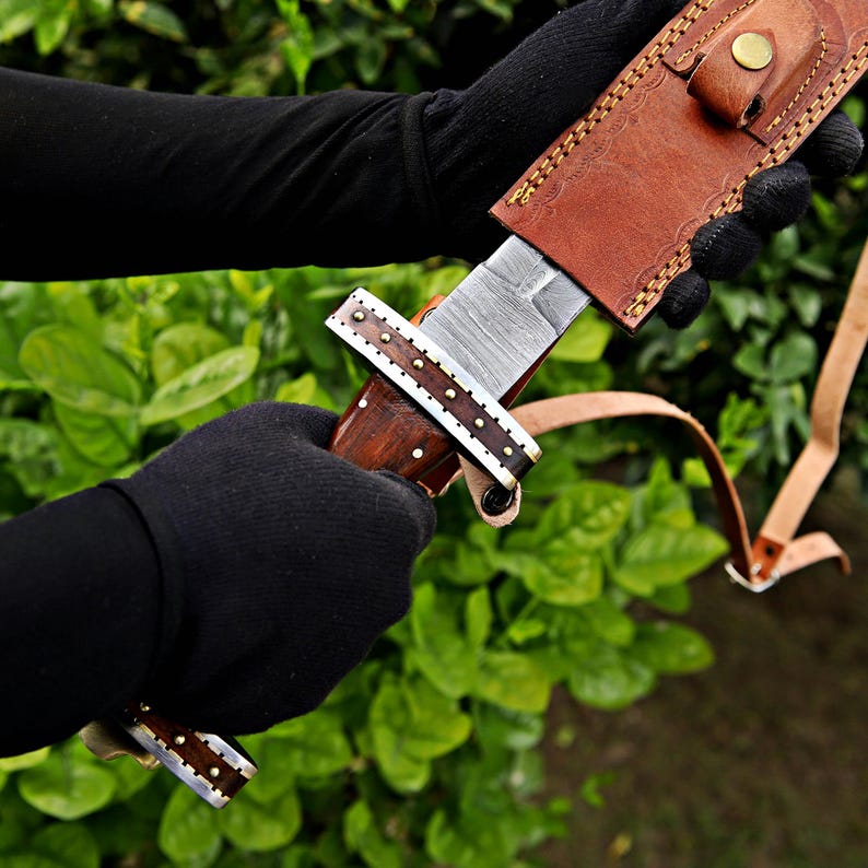 May include: A handcrafted knife with a patterned steel blade and a wooden handle. The handle is adorned with decorative metal accents. The knife is partially sheathed in a brown leather case with a strap. The background is a blurred view of green foliage.