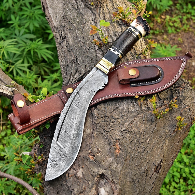 Puede incluir: Un cuchillo grande y curvo con una hoja estampada y un mango de madera oscura, apoyado en un tronco de &aacute;rbol. El cuchillo va acompa&ntilde;ado de una funda de cuero marr&oacute;n. El mango presenta detalles en lat&oacute;n y una secci&oacute;n de material claro.