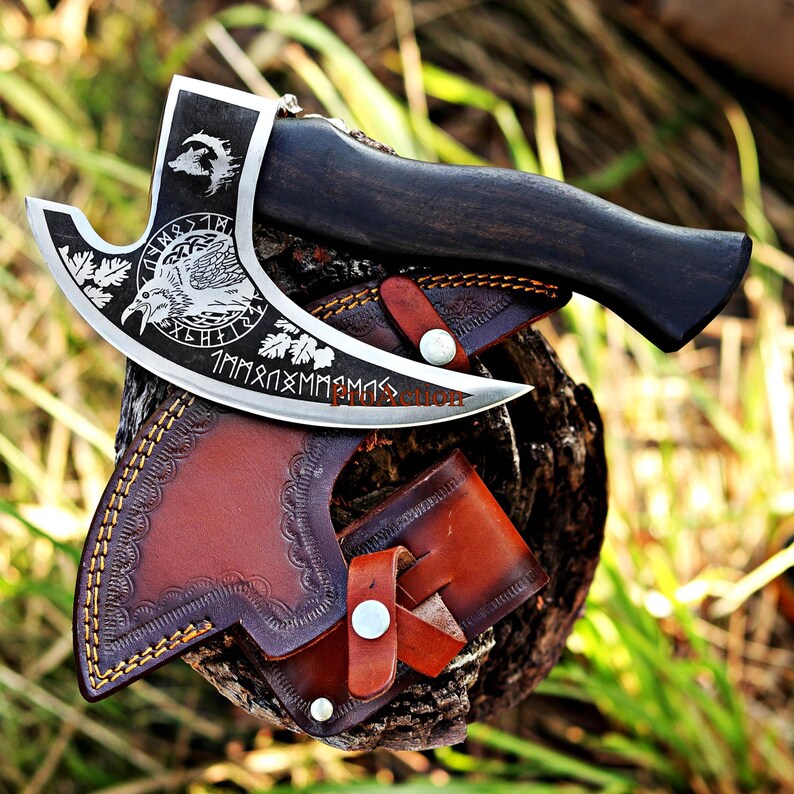 May include: A handcrafted axe with a dark wooden handle and a silver blade featuring etched designs of a wolf, a bird, and runes. The axe rests on a brown leather sheath with decorative stitching and a snap closure. The overall aesthetic is rustic and detailed.